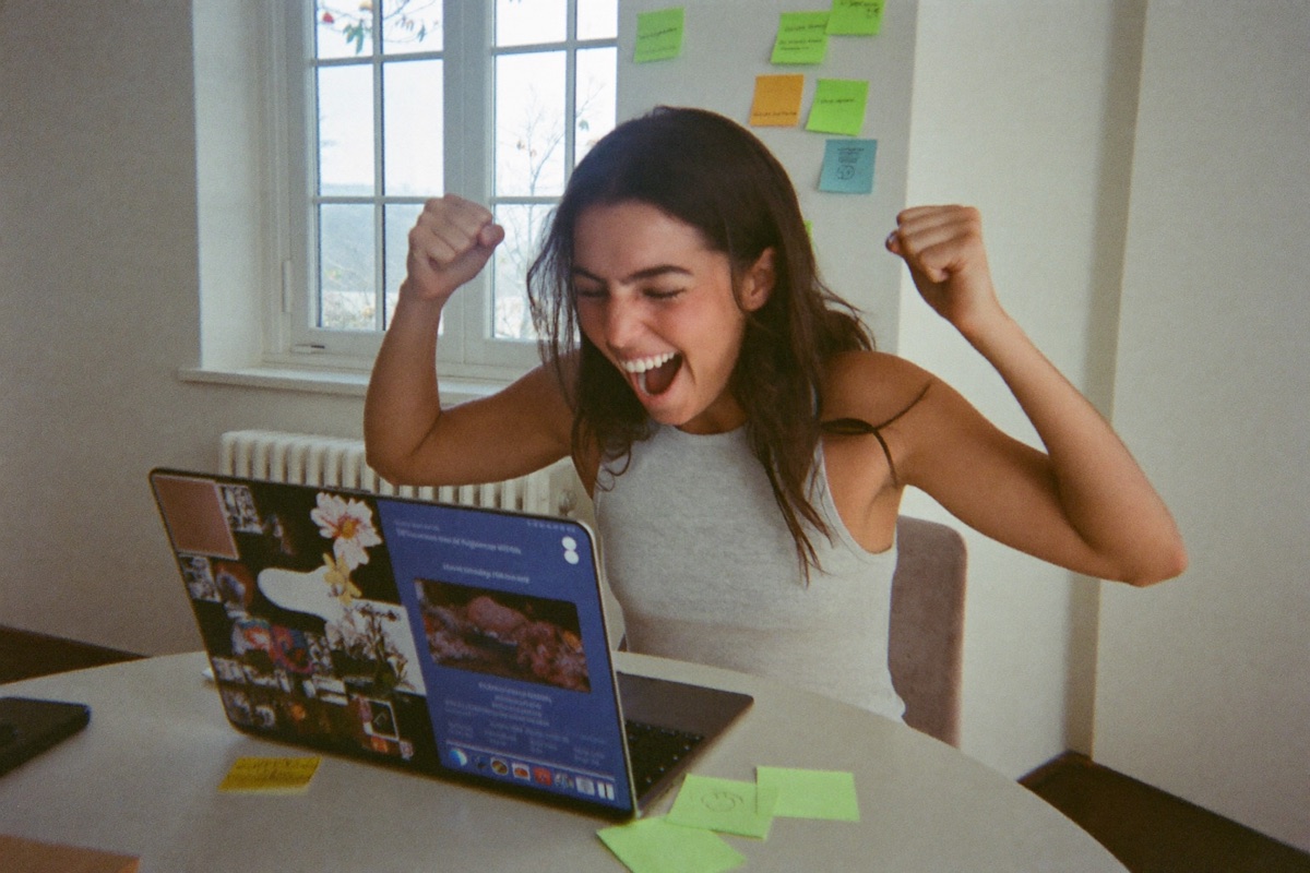 A candidate jumping up from her laptop in a bright living room with both fists raised, celebrating a passing result
