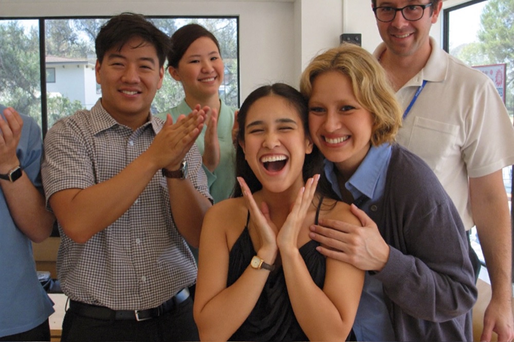 Coworkers in a bright staff room applauding and hugging a colleague celebrating her news
